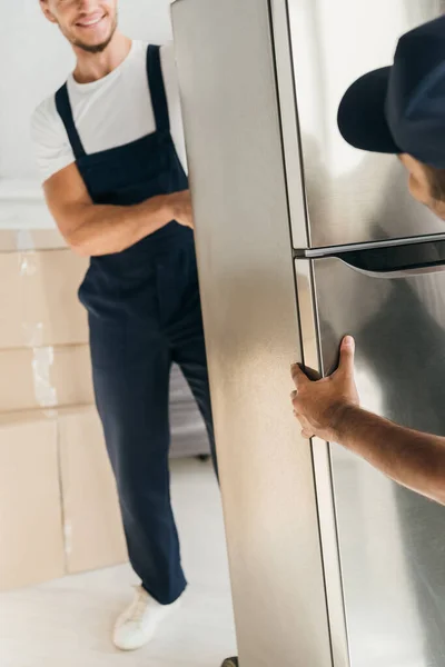 Réfrigérateur dans les mains de déménageurs multiculturels souriants en uniforme sur fond flou et au premier plan — Photo de stock