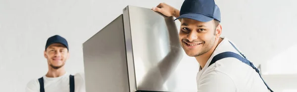 Cheerful indian mover in uniform moving fridge near coworker on blurred background, banner — Stock Photo