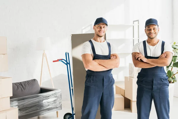 Alegres mudanzas multiculturales en gorras de pie con brazos cruzados cerca de muebles envueltos - foto de stock