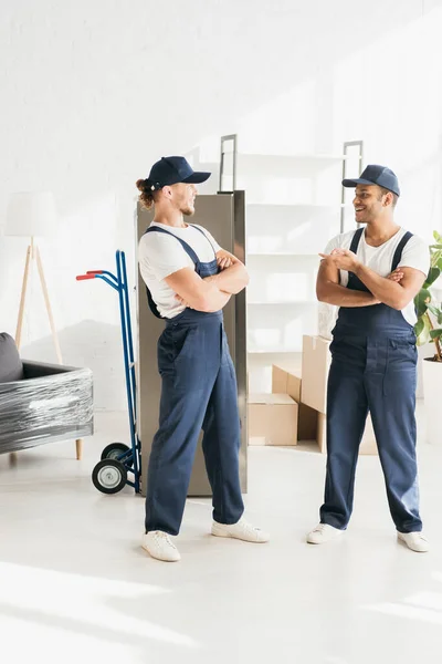 Happy indian mover pointing at coworker in cap standing with crossed arms on apartment — Stock Photo