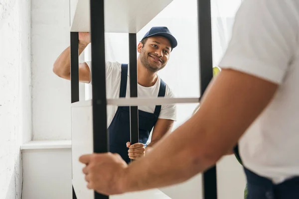 Glücklicher indischer Mover, der schweres Gestell mit Mitarbeiter auf verschwommenem Vordergrund trägt — Stockfoto