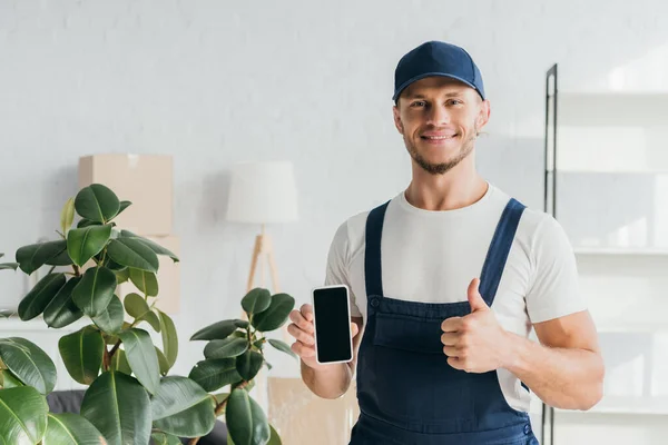 Cheerful mover in overalls holding smartphone with blanks screen and showing thumb up — Stock Photo