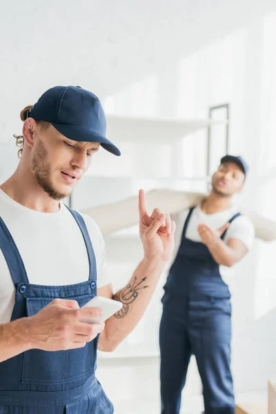 Tatuato lavoratore utilizzando smartphone e puntando con il dito, mentre mostra segno di attesa vicino indiano mover su sfondo sfocato — Foto stock