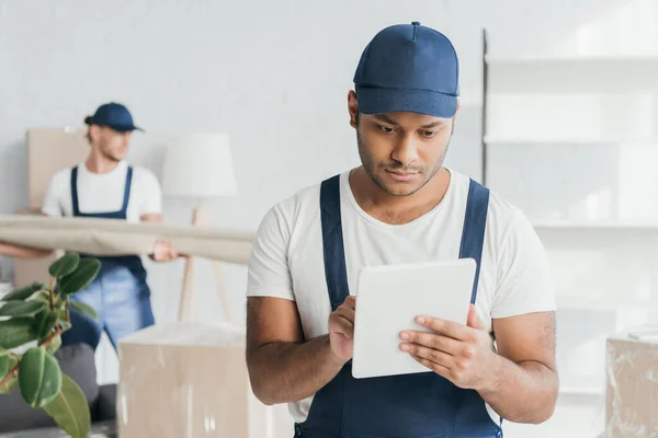 Indischer Arbeiter in Uniform mit digitalem Tablet in der Nähe von Mover hält Teppich auf verschwommenem Hintergrund — Stockfoto