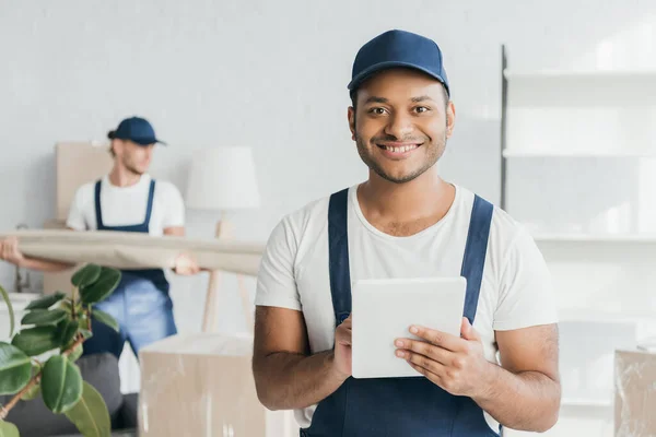 Heureux travailleur indien en uniforme en utilisant tablette numérique près mover tenant tapis sur fond flou — Photo de stock