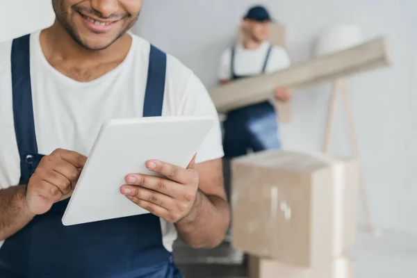 Ausgeschnittene Ansicht eines glücklichen indischen Arbeiters in Uniform mit digitalem Tablet in der Nähe des Mover auf verschwommenem Hintergrund — Stockfoto