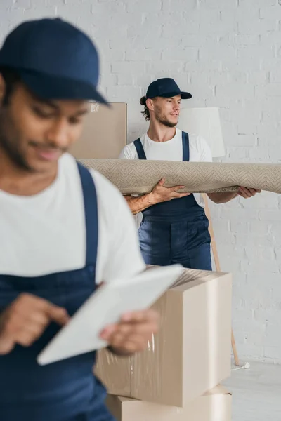 Mover holding carpet near indian worker in uniform using digital tablet on blurred foreground — Stock Photo