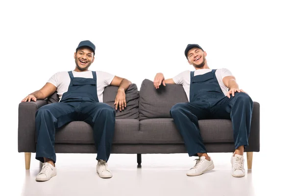 Full length of cheerful multicultural movers laughing while sitting on sofa on white — Stock Photo