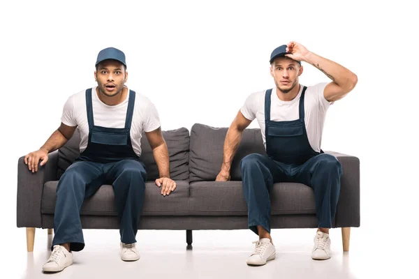 Full length of shocked mover adjusting cap while sitting on sofa with indian worker on white — Stock Photo