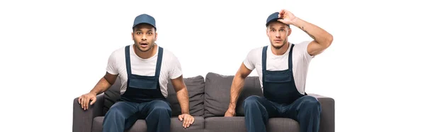 Shocked mover adjusting cap while sitting on sofa with indian worker isolated on white, banner — Stock Photo