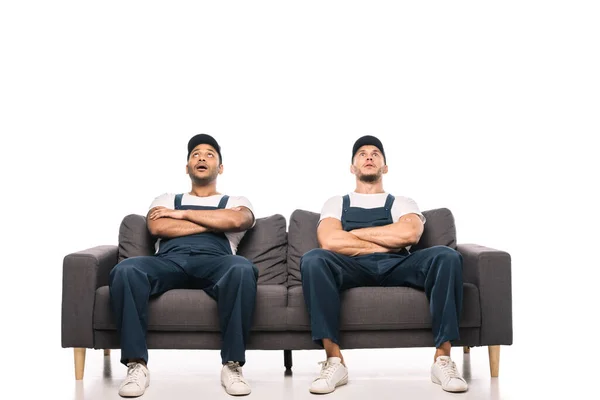 Full length of shocked multicultural movers sitting on sofa with crossed arms on while — Stock Photo