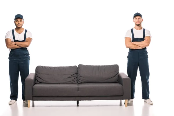 Full length of multicultural movers standing with crossed arms near couch on white — Stock Photo