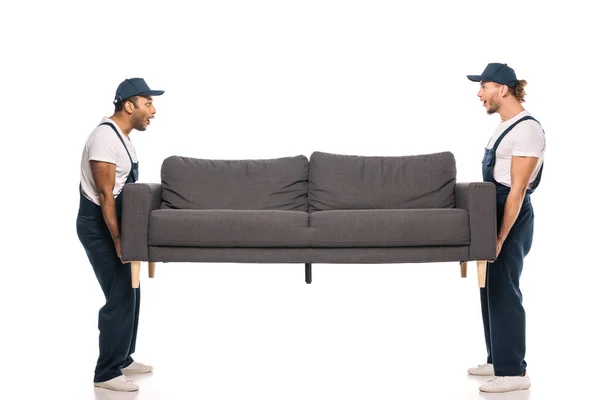 Full length of shocked multicultural movers carrying sofa on white — Stock Photo