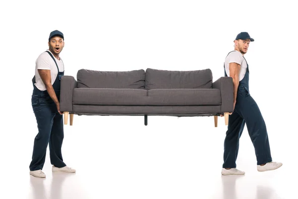 Full length of surprised multicultural movers carrying sofa on white — Stock Photo