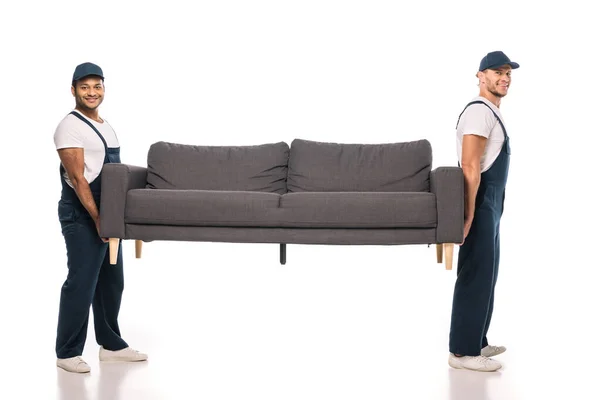 Full length of cheerful multicultural movers carrying couch on white — Stock Photo