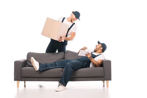 Happy indian man holding digital tablet while resting on sofa and laughing near mover with box on white — Stock Photo