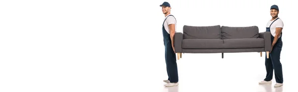 Full length of cheerful multicultural movers carrying couch on white, banner — Stock Photo