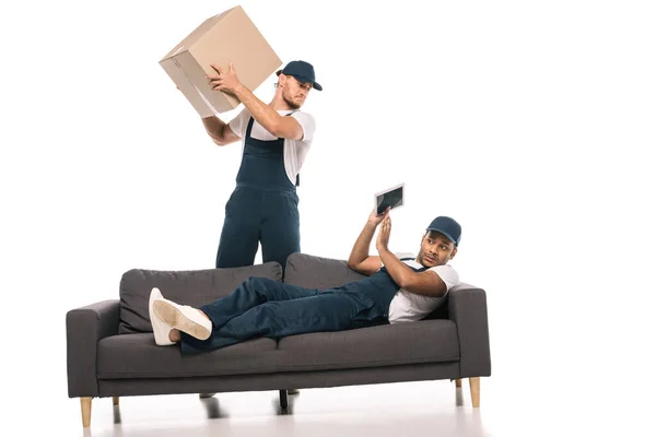 Ängstlicher indischer Arbeiter mit digitalem Tablet auf dem Sofa liegend und gestikulierend in der Nähe eines wütenden Fahrers mit Schachtel auf weißem Hintergrund — Stockfoto