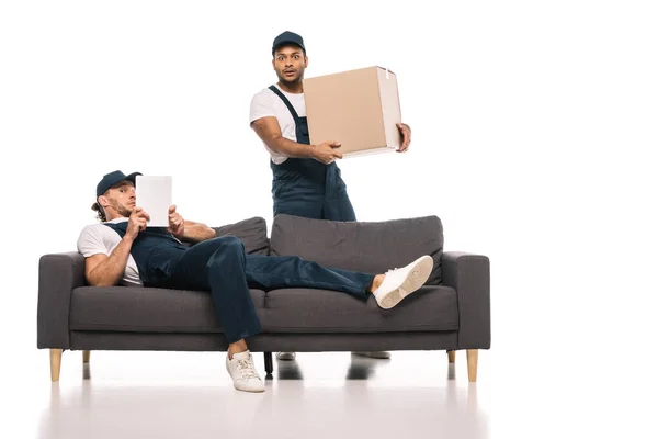 Worried worker resting on sofa with digital tablet near shocked indian mover holding box on white — Stock Photo