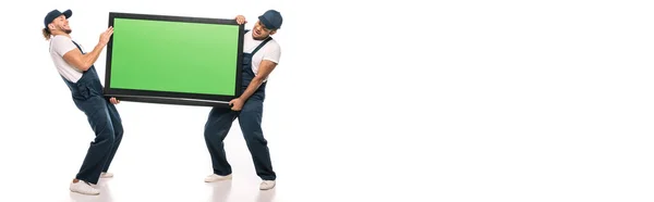 Full length of multicultural movers carrying heavy plasma tv with green screen on white, banner — Stock Photo