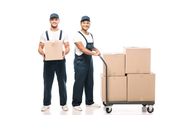 Full length of happy multicultural movers in uniform and caps near hand truck with carton boxes on white — Stock Photo