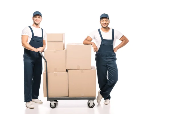 Full length of joyful multicultural movers in uniform and caps standing near hand truck with carton boxes on white — Stock Photo