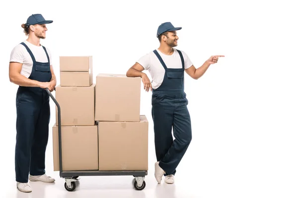 Pleine longueur de déménageur indien heureux en uniforme et chapeau pointant du doigt tout en se tenant près du camion à main avec des boîtes en carton et collègue sur blanc — Photo de stock