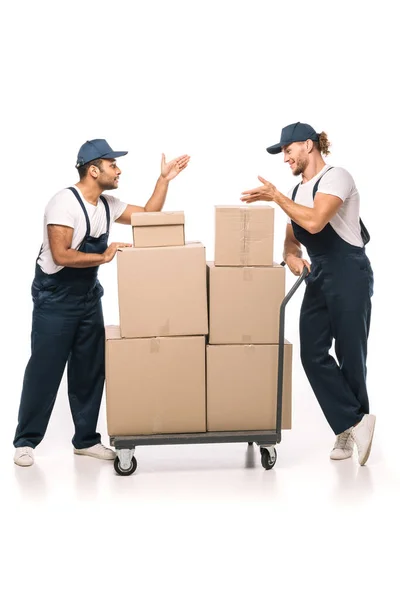 Full length of multicultural movers in uniform and caps pointing with hands at each other near hand truck with carton boxes on white — Stock Photo