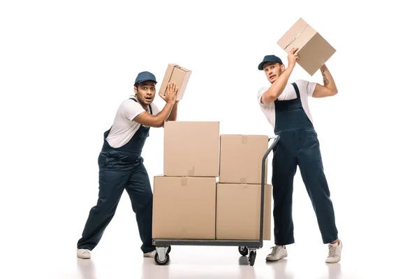 Pleine longueur de déménageurs multiculturels choqués dans des boîtes en carton de tenue uniforme près du camion à main avec des emballages sur blanc — Photo de stock