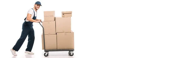 Pleine longueur de déménageur joyeux en uniforme et chapeau tirant camion à main avec boîtes en carton sur blanc, bannière — Photo de stock