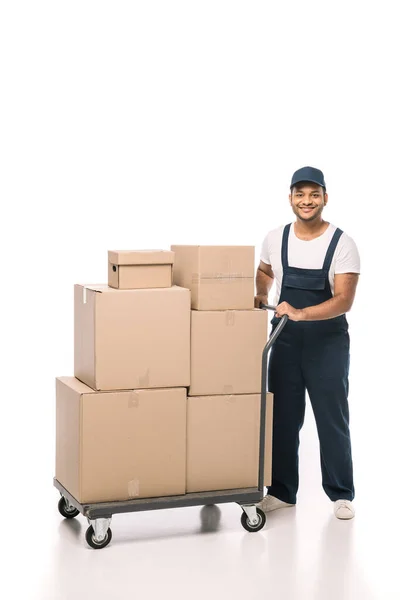 Full length of happy indian mover in overalls standing near hand truck with carton boxes on white — Stock Photo