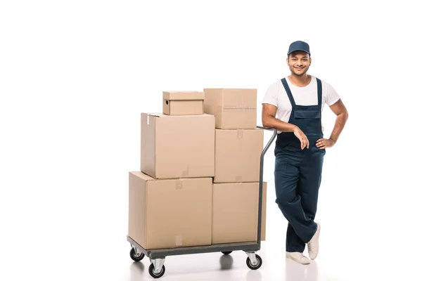 Full length of cheerful indian mover in overalls standing with hand on hip near hand truck with carton boxes on white — Stock Photo