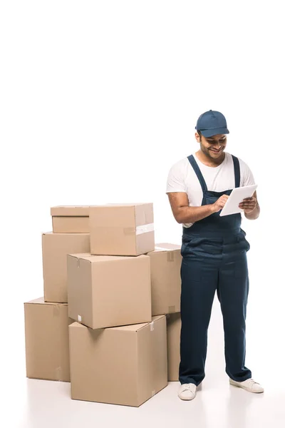 Happy indian mover in overalls using digital tablet near carton boxes on white — Stock Photo