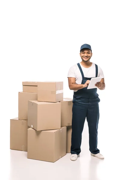 Joyeux déménageur indien en salopette en utilisant une tablette numérique près de boîtes en carton sur blanc — Photo de stock