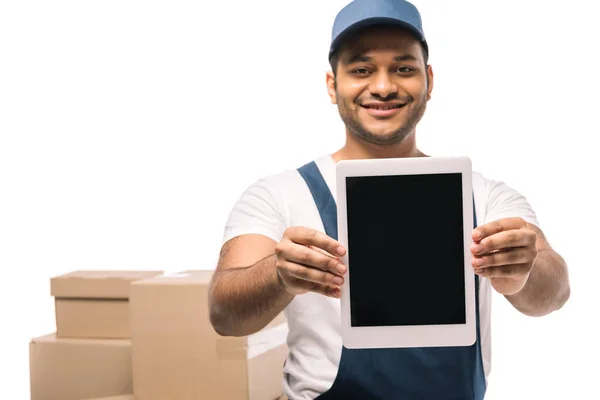 Feliz indio mover en overoles celebración tableta digital con pantalla en blanco cerca de cajas de cartón sobre fondo borroso aislado en blanco - foto de stock