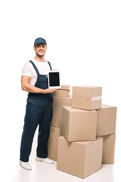 Full length of cheerful mover in uniform and cap holding digital tablet with blank screen near carton boxes on white — Stock Photo