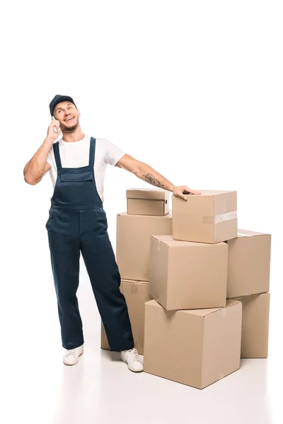 Full length of cheerful mover in uniform and cap talking on smartphone near carton boxes on white — Stock Photo