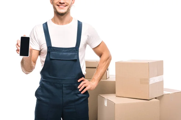 Vista parcial de alegre mover en uniforme celebración de teléfono inteligente con pantalla en blanco cerca de cajas de cartón aislado en blanco - foto de stock