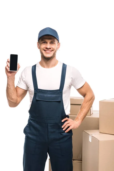 Cheerful mover in uniform holding smartphone with blank screen and standing with hand on hip near carton boxes isolated on white — Stock Photo