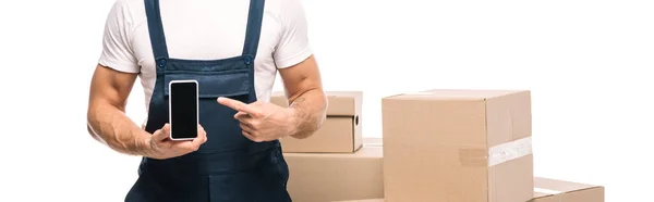 Cropped view of mover in uniform pointing with finger at smartphone near carton boxes isolated on white, banner — Stock Photo