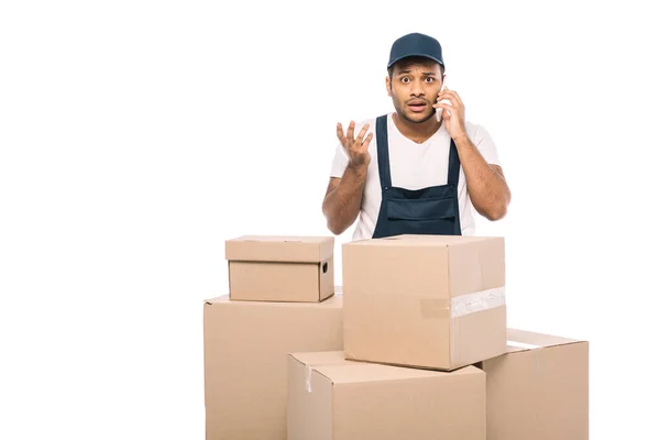 Displeased indian mover in overalls talking on smartphone near carton boxes isolated on white — Stock Photo