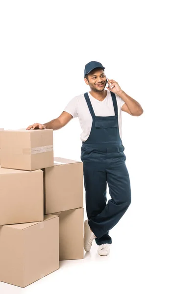 Full length of happy indian mover in overalls talking on smartphone while standing near carton boxes on white — Stock Photo