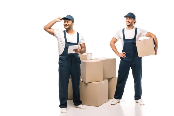 Full length of cheerful indian worker holding digital tablet, adjusting cap and standing with mover near boxes on white — Stock Photo