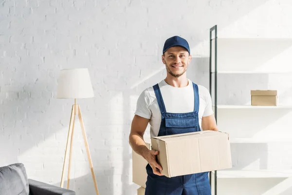 Alegre mover em cap segurando caixa de papelão no apartamento — Fotografia de Stock