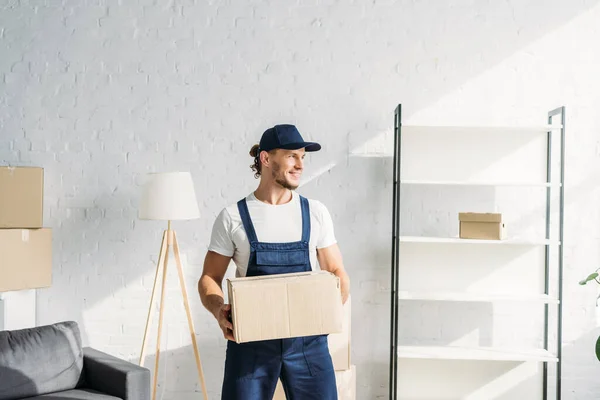 Happy mover in cap holding carton box and looking away in apartment — Stock Photo