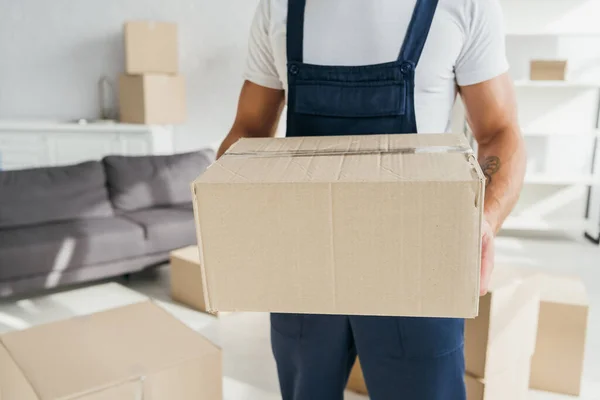 Cropped view of tattooed mover holding carton box in apartment — Stock Photo