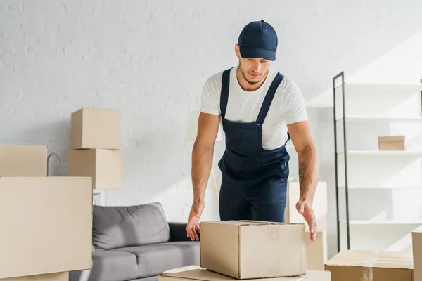 Tattooed mover in overalls and cap reaching box in apartment — Stock Photo