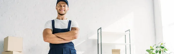 Happy mover in uniform and cap standing with crossed arms in apartment, banner — Stock Photo