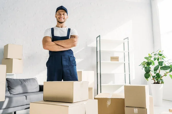 Cheerful mover in uniform and cap standing with crossed arms near boxes in apartment — Stock Photo