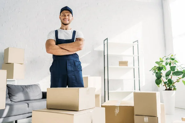 Allegro traslocatore in uniforme e cappuccio in piedi con braccia incrociate vicino scatole in appartamento — Foto stock